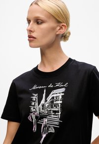 Blonde woman with hair in a bun wearing a black T-shirt featuring a white and pink sketch of Paris street and Eiffel Tower.