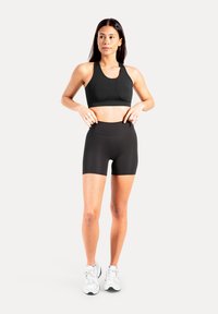 Svart sport-bh och svart högmidjade shorts. Slätt tyg, figursydd design med sömlösa kanter. Modellen bär vita sneakers.