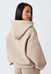 Sudadera beige con capucha de corte holgado, que presenta una textura de tela suave, capucha grande y hombros caídos. Diseño sencillo sin estampados.