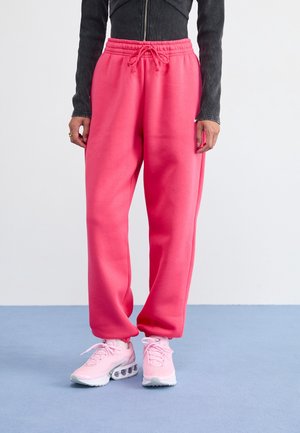 Personne portant un pantalon de survêtement rose vif avec cordon de serrage et des chaussures de sport rose clair, debout devant un fond uni.