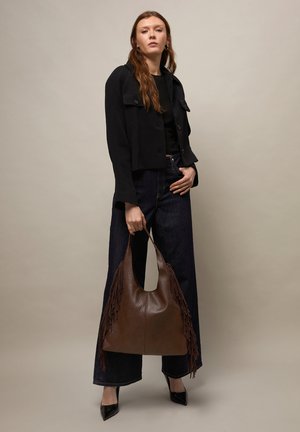 Frau trägt schwarze kurze Jacke und weit geschnittene Jeans, hält große braune Leder-Handtasche mit Fransen, steht vor einfarbigem beigem Hintergrund.