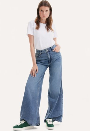 Jeans in denim blu a gamba larga con vita alta, abbinati a una maglietta bianca semplice e scarpe da ginnastica verdi. Tessuto testurizzato con cuciture visibili.