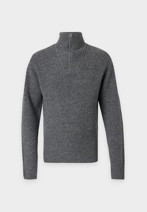 Un jeune homme porte un pull vert foncé à demi-fermeture éclair sur une chemise bleue, un pantalon bleu marine avec une ceinture noire et des bottes en cuir noires.