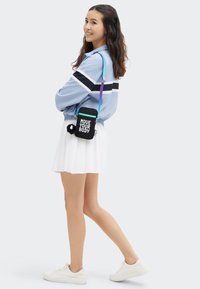 Borsa a tracolla nera con testo "MUOVI IL TUO CORPO", zipper turchese e pom-pom nero rotondo, indossata con una giacca sportiva blu e una gonna bianca.