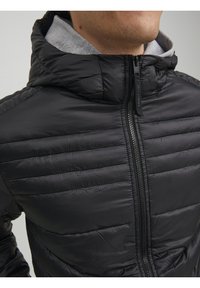 Hombre con una chaqueta acolchada negra con capucha y un forro interior gris, cremallera completamente cerrada hasta el cuello.