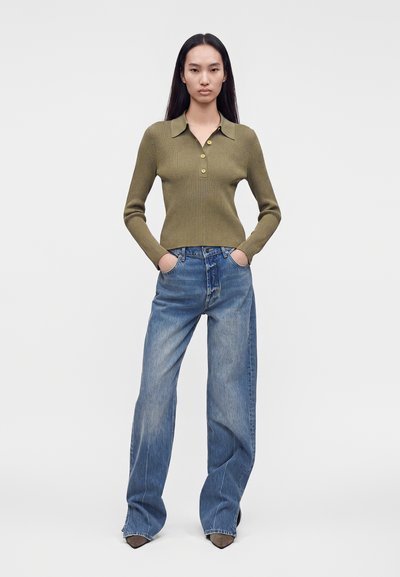 Femme aux longs cheveux noirs portant un pull côtelé vert olive et un jean bleu à jambes larges, les mains dans les poches, sur un fond uni.