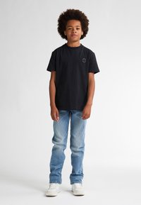 Zwarte katoenen t-shirt met een rond logo op de borst, gecombineerd met lichtblauwe spijkerbroek en witte sneakers. Eenvoudige achtergrond.