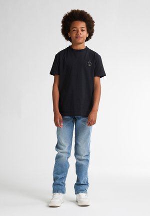 Zwarte katoenen t-shirt met een rond logo op de borst, gecombineerd met lichtblauwe spijkerbroek en witte sneakers. Eenvoudige achtergrond.