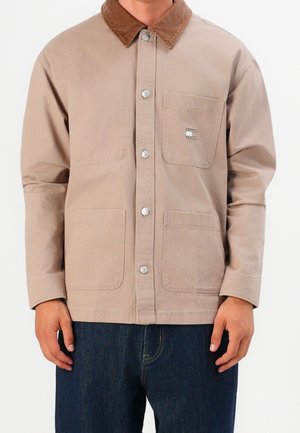 Beige skjortejakke med brun kordfløjls-krave, tre forlommer og Tommy Jeans-logo på brystlommen, båret med mørkeblå jeans.