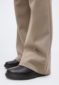 Schwarze Lederschuhe, getragen zu beigen Hosen mit "J.LINDEBERG" Stickerei nahe dem Saum, vor einem schlichten weißen Hintergrund.