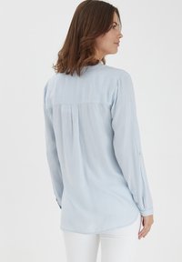Light blue striped long-sleeve shirt with a back pleat, buttoned cuffs, and a rounded hem, made from a soft fabric.