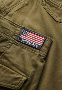 Olijfgroen stof met een textuur, voorzien van een geborduurd patch met een kleine Amerikaanse vlag en "SUPERDRY" in het wit.
