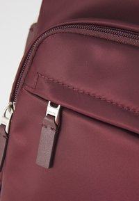 Bordový ruksak z hladkej nylonovej tkaniny, ktorý má zipsovú kapsu a kovový ťahák s nápisom "Calvin Klein."