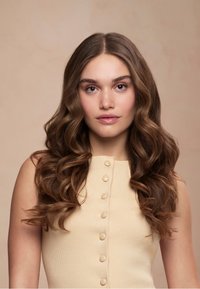 Jeune femme aux longs cheveux bruns ondulés, portant un haut beige sans manches avec des boutons, regardant directement la caméra avec une expression neutre.