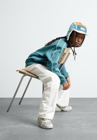 Teal sweatshirt with white accents, white pants, and gray sneakers. A multicolored hat sits atop braided hair, while seated on a metal chair.