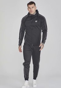 Fekete tréningruha cipzáras kapucnival, húzózsinórral, fényvisszaverő csíkokkal és hozzáillő nadrággal. Fehér tornacipővel viselt.