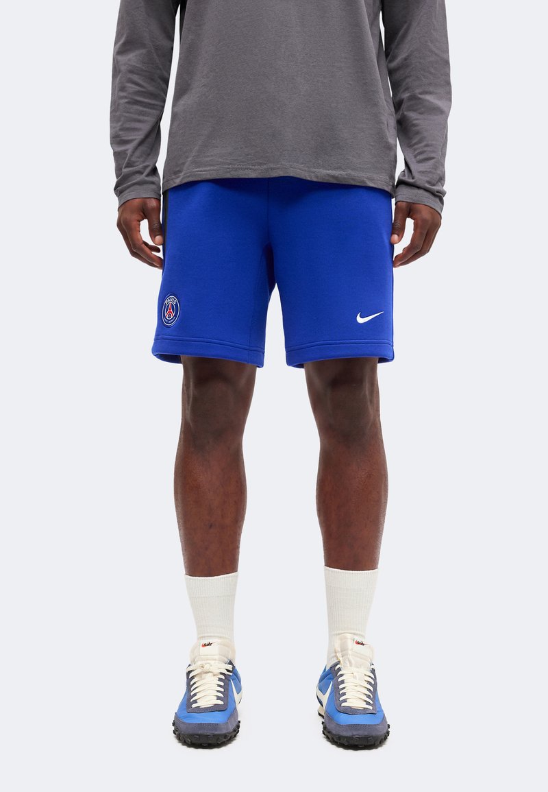 Άνδρας που φορά μπλε σορτς Paris Saint-Germain Nike, γκρι μακρυμάνικη μπλούζα, λευκές κάλτσες και μπλε αθλητικά παπούτσια Nike, στέκεται μπροστά σε απλό φόντο.