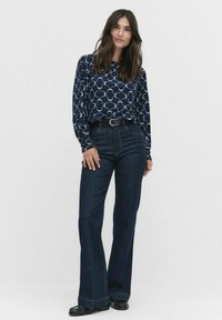 Marinblå långärmad topp med vitt geometriskt mönster, kombinerad med mörk tvättade utsvängda jeans. Svart bälte med silverfärgad spänne och svarta skor.