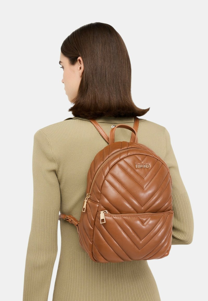 Zaino in pelle trapuntata marrone con manico superiore, spallacci regolabili e tasca frontale con zip. Presenta un dettaglio logo in rilievo.