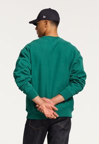 Felpa verde in cotone con polsini e orlo a coste. Modello visto da dietro, indossa un cappellino blu navy. Jeans scuri visibili in vita.