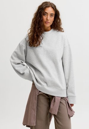 Kobieta z kręconymi włosami, ubrana w jasnoszary sweatshirt, beżowe spodnie i czerwoną koszulę w kratę zawiązaną w talii na jednolitym tle.