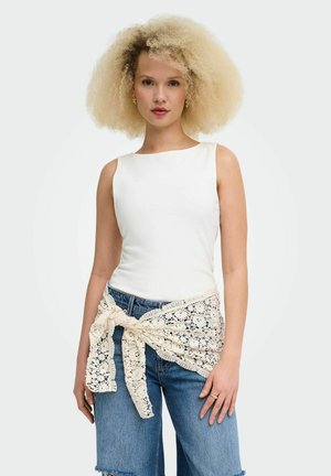 Giovane donna con capelli biondi e ricci che indossa un top bianco senza maniche, jeans blu e uno scialle beige in pizzo annodato in vita.