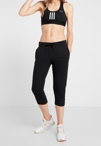 Sujetador deportivo negro con tres rayas blancas en la parte frontal, complementado con pantalones de chándal cortos negros y zapatillas deportivas de color claro.
