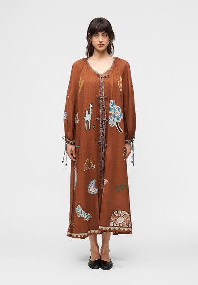 Femme portant une robe longue ample de couleur rouille avec des imprimés abstraits colorés, manches longues avec cordons, et chaussures plates noires, debout devant un fond blanc.