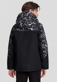 Chłopiec tyłem, ubrany w czarną kurtkę z białym marmurkowym wzorem na kapturze i rękawach, na jednolitym jasnoszarym tle.