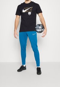 T-shirt noir Nike avec logo doré, pantalon de sport bleu avec accents latéraux et chaussures de foot noires tenant un ballon Nike F.C. noir et gris.