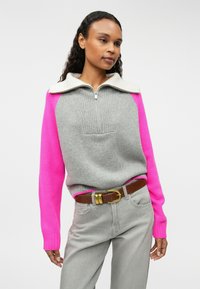 Pull color bloc gris et rose avec un col zippé, texture côtelée sur la poitrine, contrastée par des manches roses lisses. Associé à un pantalon gris.