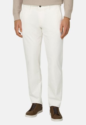 Mann trägt weiße, gerade geschnittene Hosen, beige Pullover und braune Sneakers mit weißen Sohlen, steht vor einem einfarbigen Hintergrund.