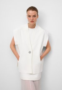Débardeur blanc sans manches à coupe ample, avec une fermeture à l'avant dotée d'un bouton argenté, superposé sur un haut blanc court et une jupe plissée transparente.