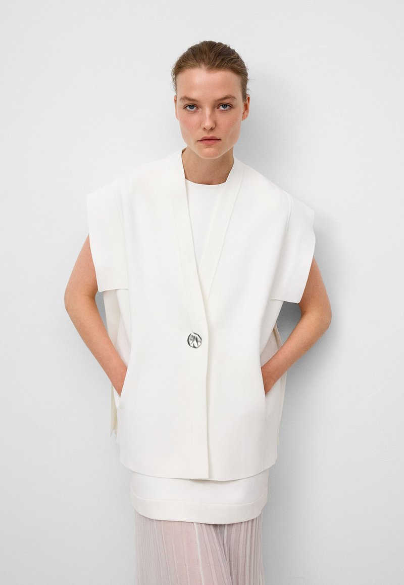 Débardeur blanc sans manches à coupe ample, avec une fermeture à l'avant dotée d'un bouton argenté, superposé sur un haut blanc court et une jupe plissée transparente.