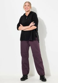 Zwart, lang mouwens hoodie met ritssluiting, gecombineerd met paarse, elastische taillebroek en zwarte laarzen. De outfit heeft een relaxte pasvorm.