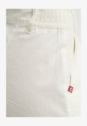 Śmietankowe spodnie dresowe z elastyczną talią i boczną kieszenią, z małą czerwoną metką z logo w kształcie gwiazdy na prawej kieszeni.