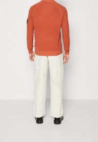 Rostfärgad tröja med ribbad struktur, raglanärmar och bandade muddarna, i kombination med ljusbeiga cargobyxor och svarta skor.