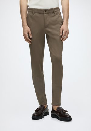 Homme portant un pantalon marron slim associé à des mocassins à glands marron foncé sur un fond clair uni.