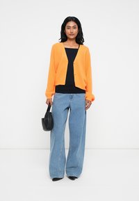 Oranža adīta kardigāna ar brīvu piegriezumu, balti pogas, valkāta virs melna topa, saskaņota ar platām zilu džinsu biksēm un melnu rokassomu.