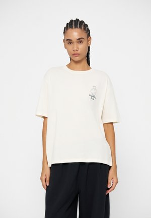 Jeune femme aux cheveux tressés portant un t-shirt blanc ample avec un petit vase et le texte "Ceramics Club", associé à un pantalon noir à jambes larges.