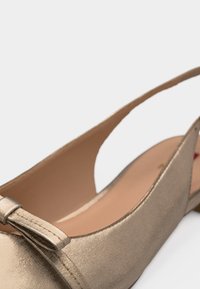 Chaussure plate pour femmes couleur or avec un petit nœud sur l'avant et un design à talon ouvert.