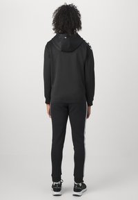 Persona de pie de espaldas con sudadera negra con capucha, pantalones deportivos negros con franjas blancas laterales y zapatillas negras sobre fondo blanco.