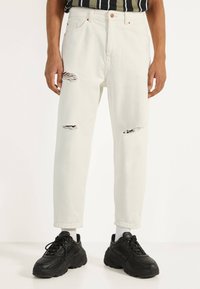 Bílé džíny z denimu s roztrhanými detaily na kolenou, rovný střih, vysoký pas a měděné doplňky. Kombinovány s černými robustními teniskami.