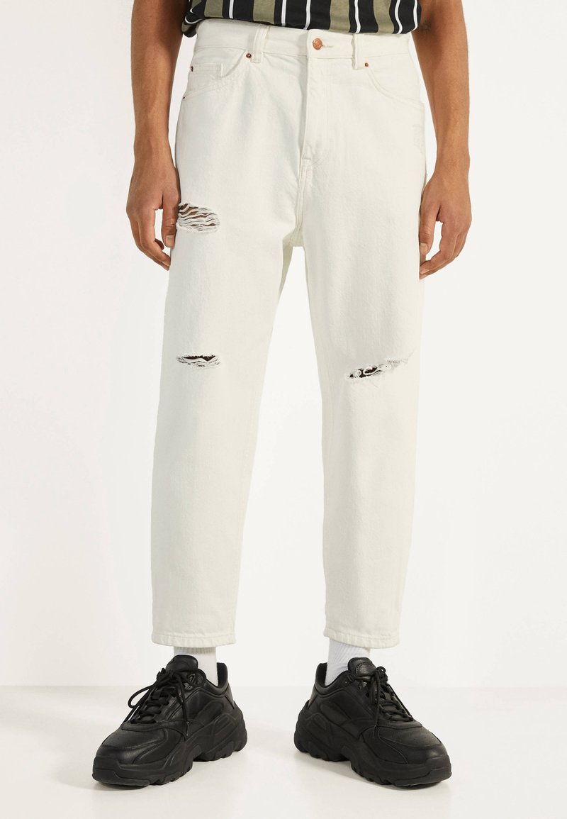 Bílé džíny z denimu s roztrhanými detaily na kolenou, rovný střih, vysoký pas a měděné doplňky. Kombinovány s černými robustními teniskami.