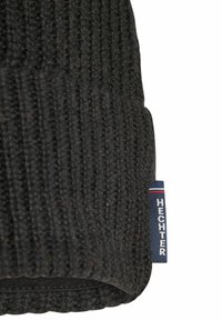Zwarte gebreide muts met geribbelde textuur. Voorzien van een klein blauw label met de tekst "HECHTER" en rood-witte accenten. Eenvoudig, functioneel ontwerp.