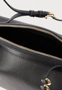 Sort læderhåndtaske med lynlås i guldtonet metal, tekstureret overflade og en flad strop med metalpynt. Indvendigt er foret mørkt.