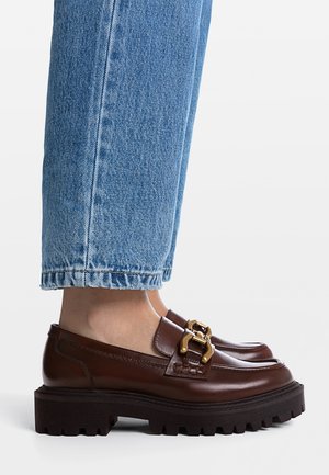 Mocassins en cuir marron avec une semelle noire épaisse et des détails en or, associés à un jean bleu retroussé montrant un ourlet effiloché.