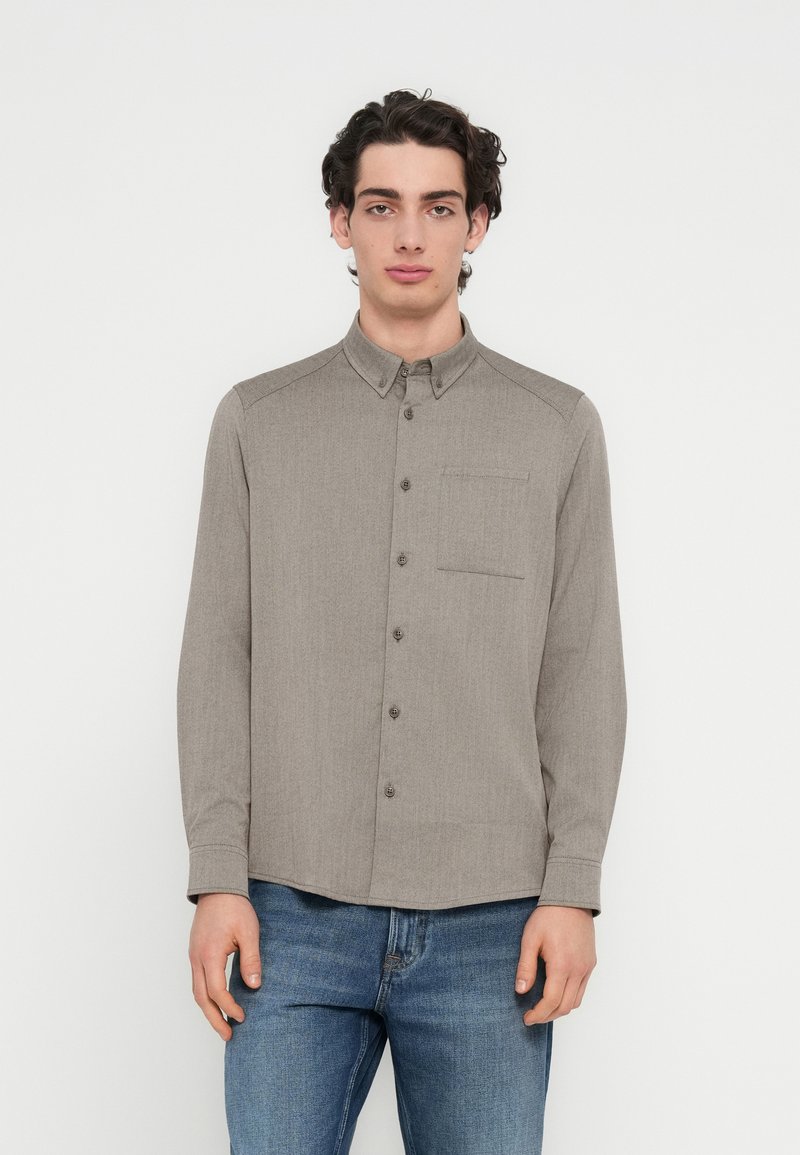 Chemise grise à boutons avec un motif subtil, manches longues et poche poitrine. Fabriquée dans un tissu texturé. Portée avec un jean bleu.