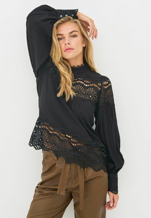 Svart blus med spetsdetaljer längs fållen och manschetterna, med hög krage och svängda ärmar. Bärs med bruna byxor.