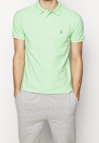 Muž v světle zeleném polo tričku s krátkým rukávem a malým vyšitým logem, světlé šedé tepláky, ruce v kapsách.